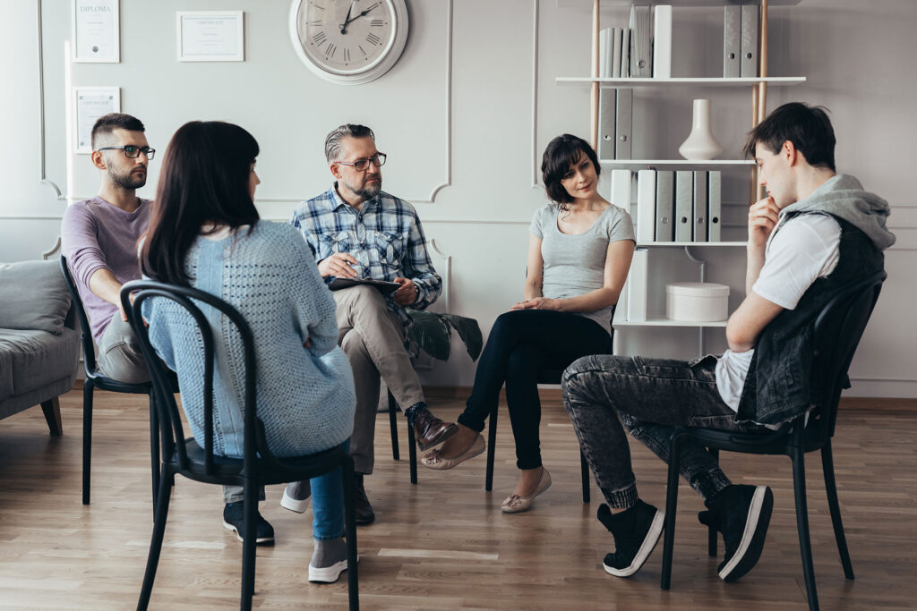 Five People Seated In Chairs, Conversing In A Well-Lit Room With Neutral Walls